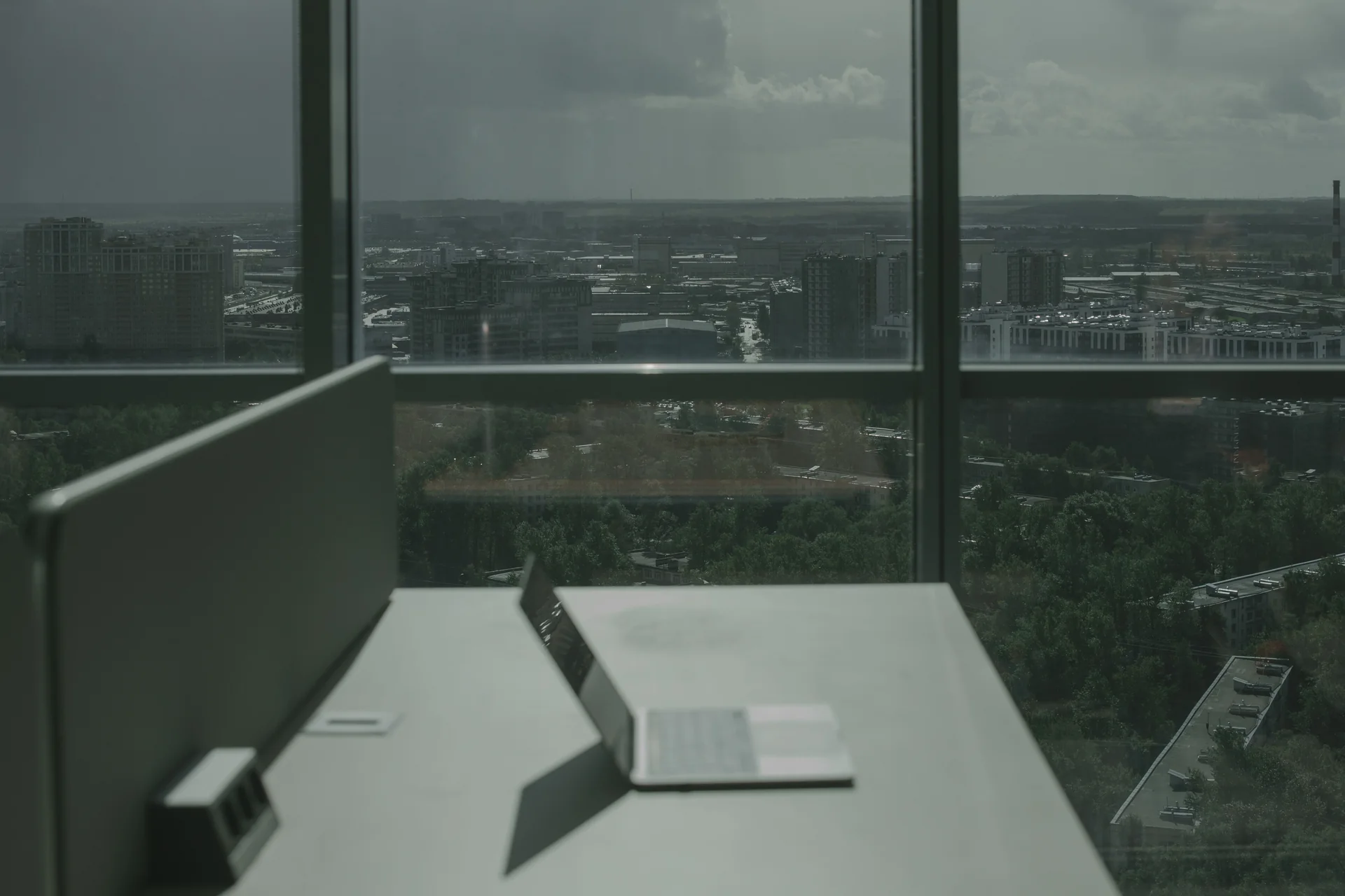 An open laptop on a desk beside a floor-to-ceiling window overlooking a city.